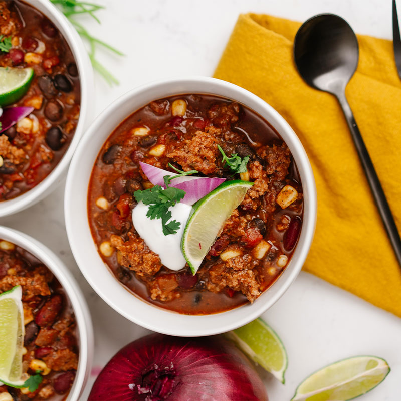Bowl of taco soup with toppings including lime, onion, and sour cream on a white surface with a yellow napkin.
