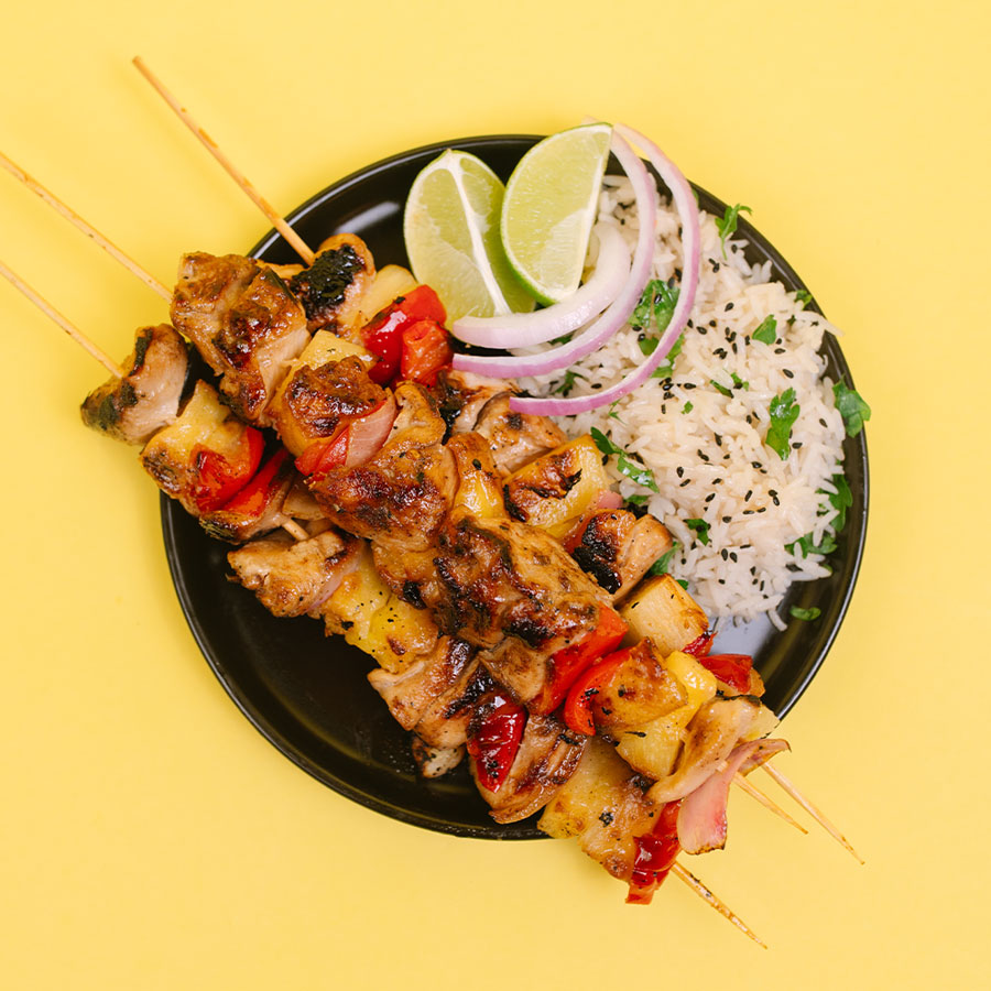 Grilled chicken and pineapple skewers served on a black plate with white rice, sliced red onion, lime wedges, and black sesame seeds, set against a solid yellow background.