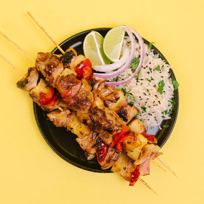 Grilled chicken and pineapple skewers served on a black plate with white rice, sliced red onion, lime wedges, and black sesame seeds, set against a solid yellow background.