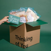 A cardboard gift box printed with the words 'Thinking of You' sits open on a dark green background. A hand on the left holds a matching teal card inside the box, which is filled with packaged food items.