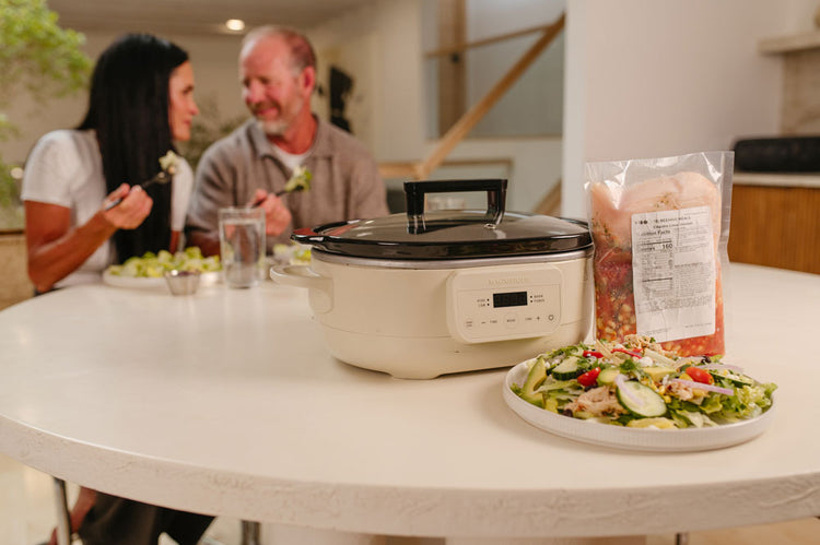 A cream-colored slow cooker, a frozen Beehive Meal, and a salad sits on a white dining table in a home kitchen setting. People are in the background eating dinner. 