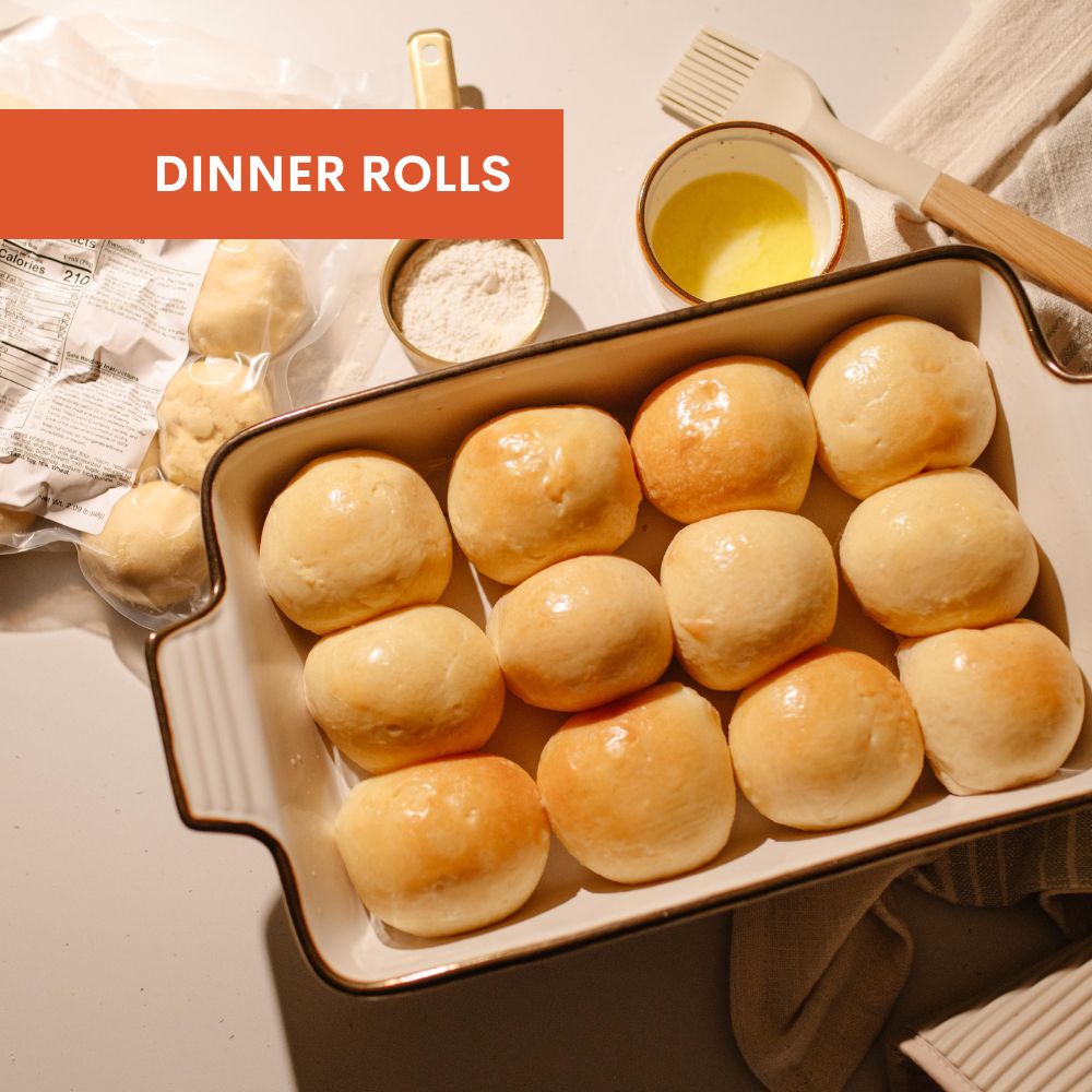 Baked dinner rolls in a baking dish with a red label, surrounded by flour, butter, and a brush.