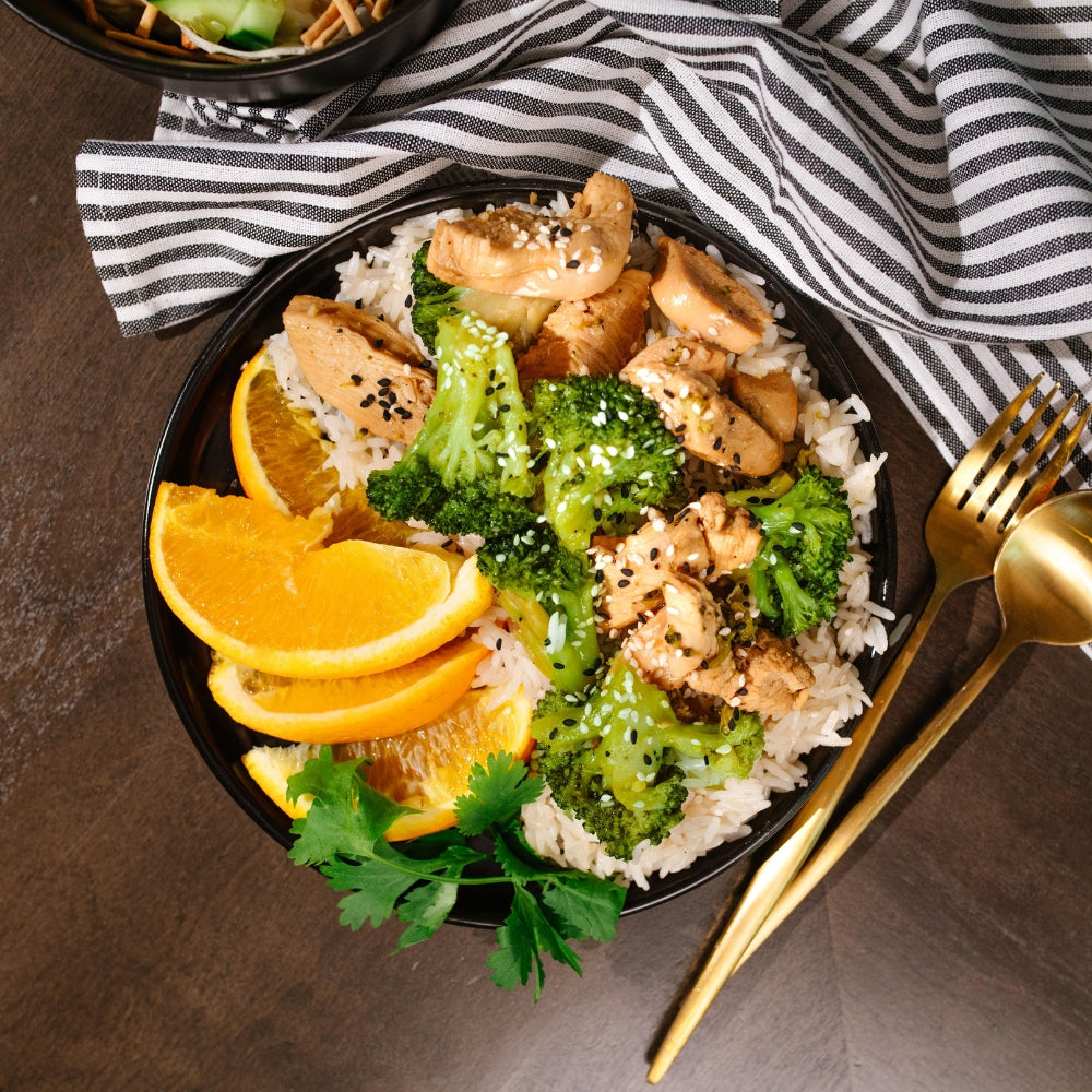 Bowl of Beehive Meals Orange Chicken with broccoli and oranges on a plate next to a striped cloth