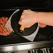Person using a tool to press smash burgers on a grill plate with a close-up view.