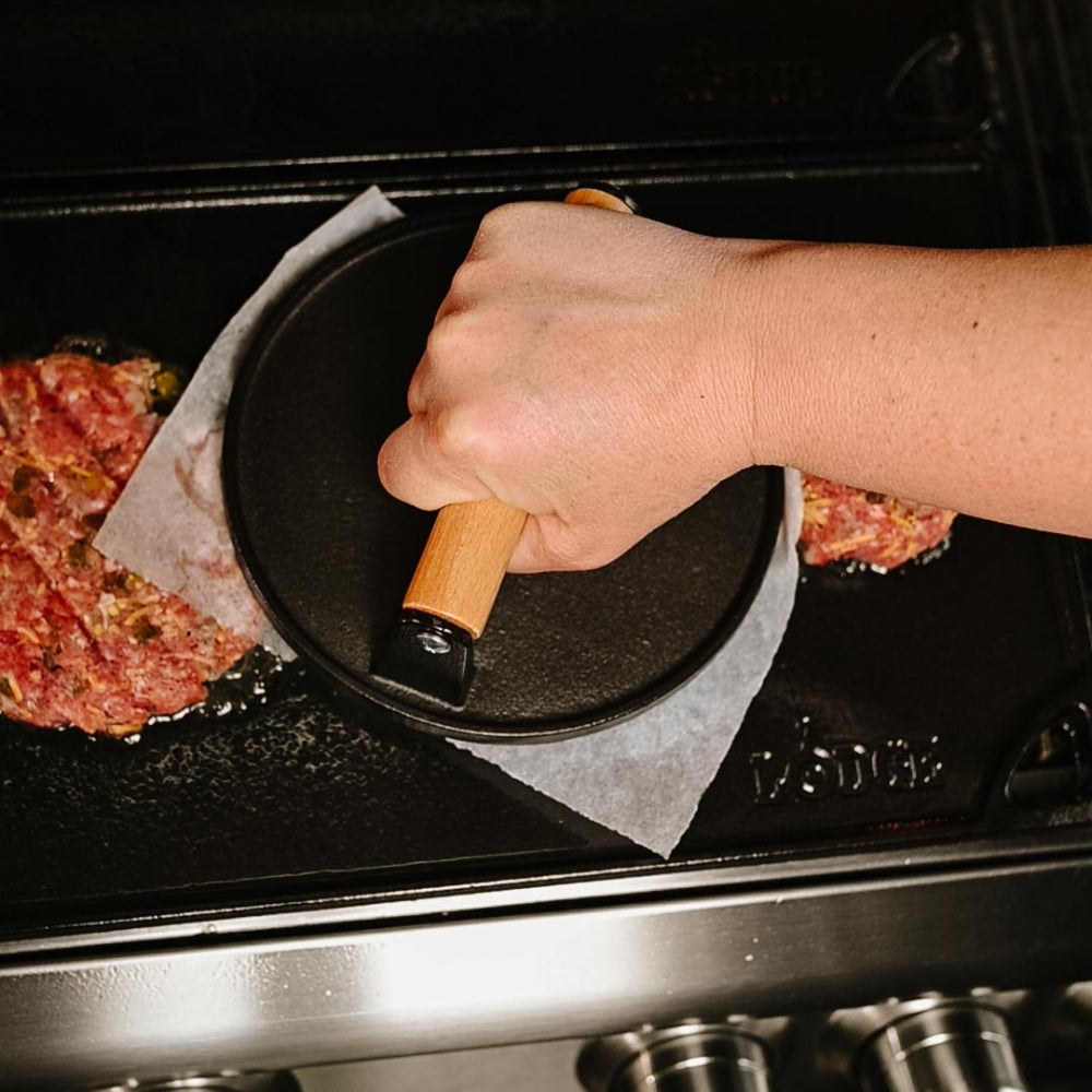 Person using a tool to press smash burgers on a grill plate with a close-up view.