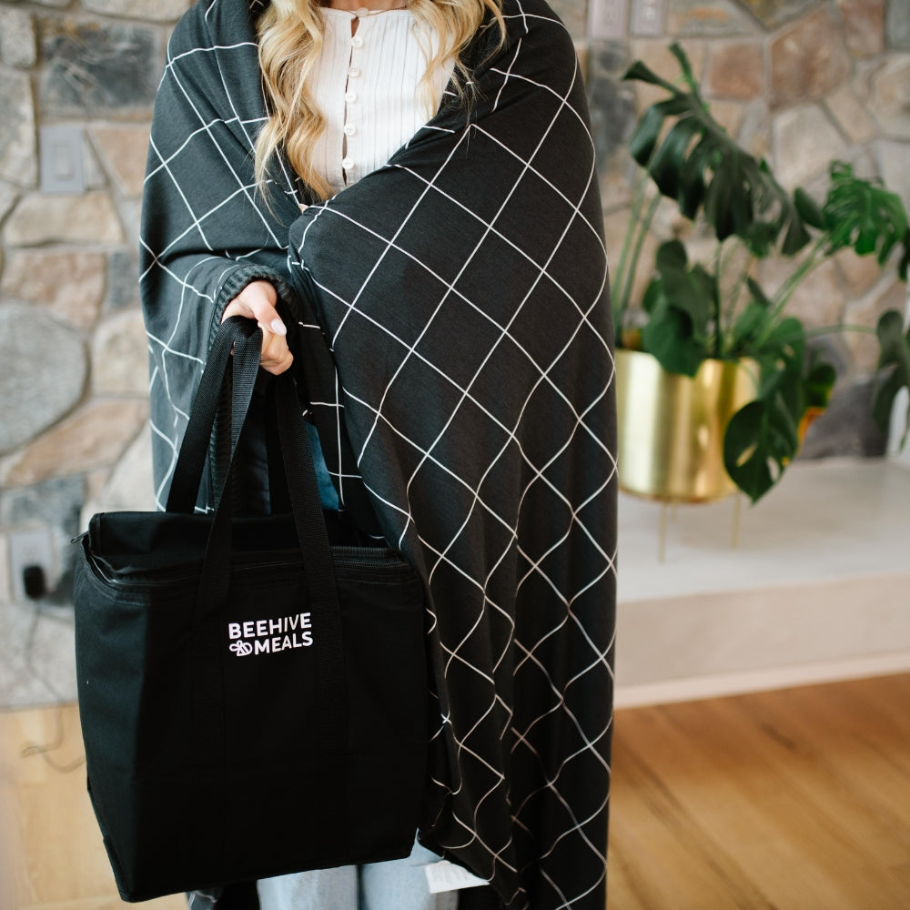 Person wrapped in a black and white grid-patterned blanket holding a black tote bag with 'Beehive Meals' logo.