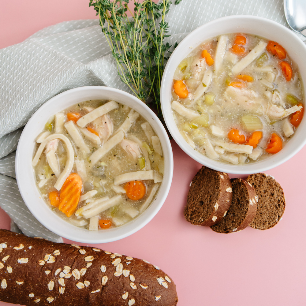 Two bowls of chicken noodle soup with carrots and bread on a pink background