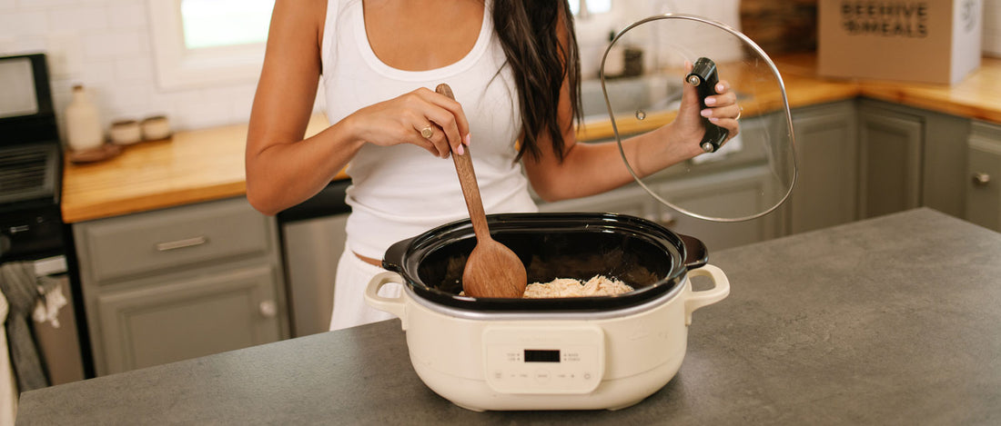 A woman in a white tank top stirs Beehive Meals food inside a cream-colored slow cooker on a kitchen counter, with her glass lid propped beside it in a home kitchen setting.