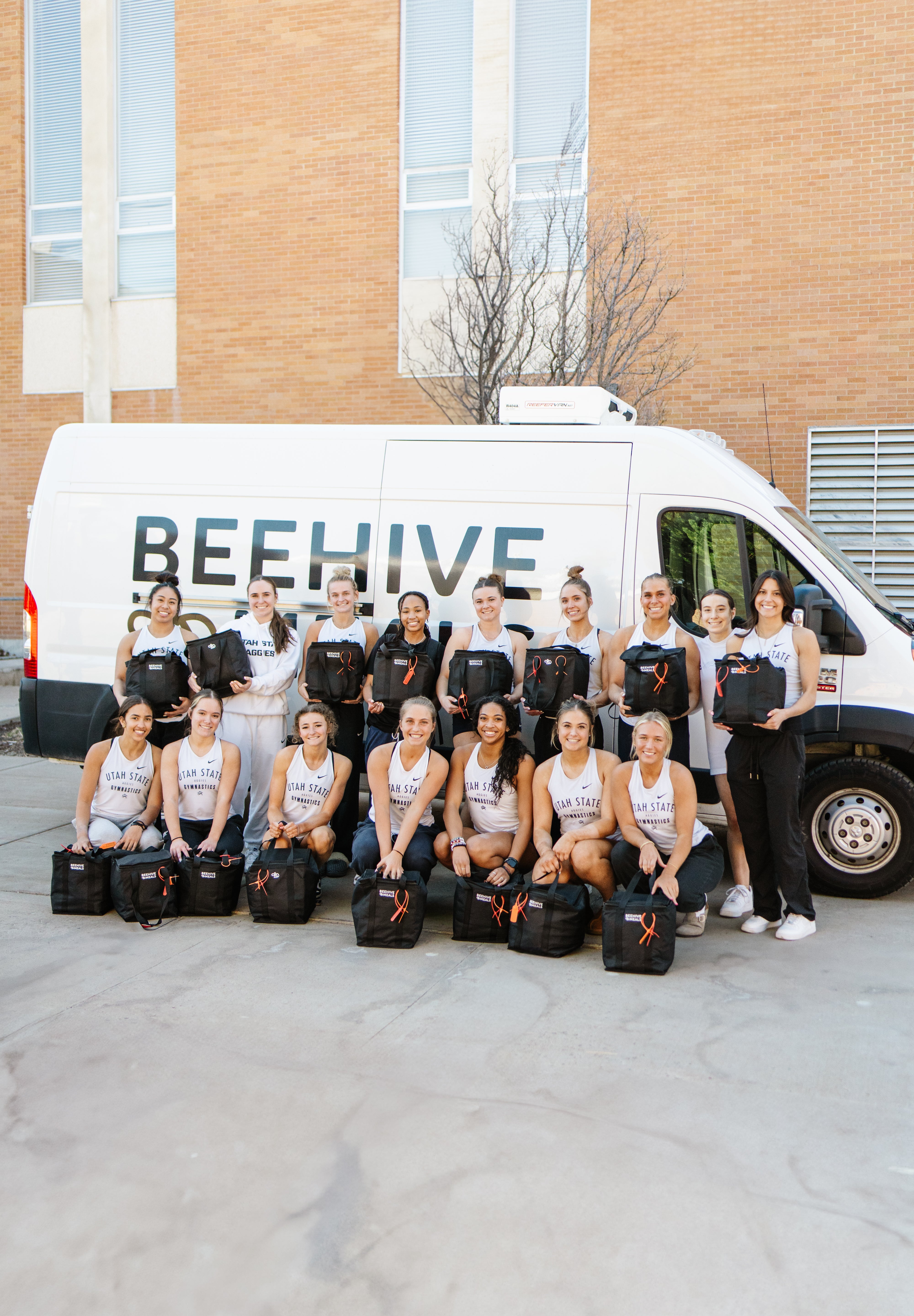 The 2025 Utah State Gymnastics team posing with a delivery of Beehive Meals in front of a Beehive Meals van.