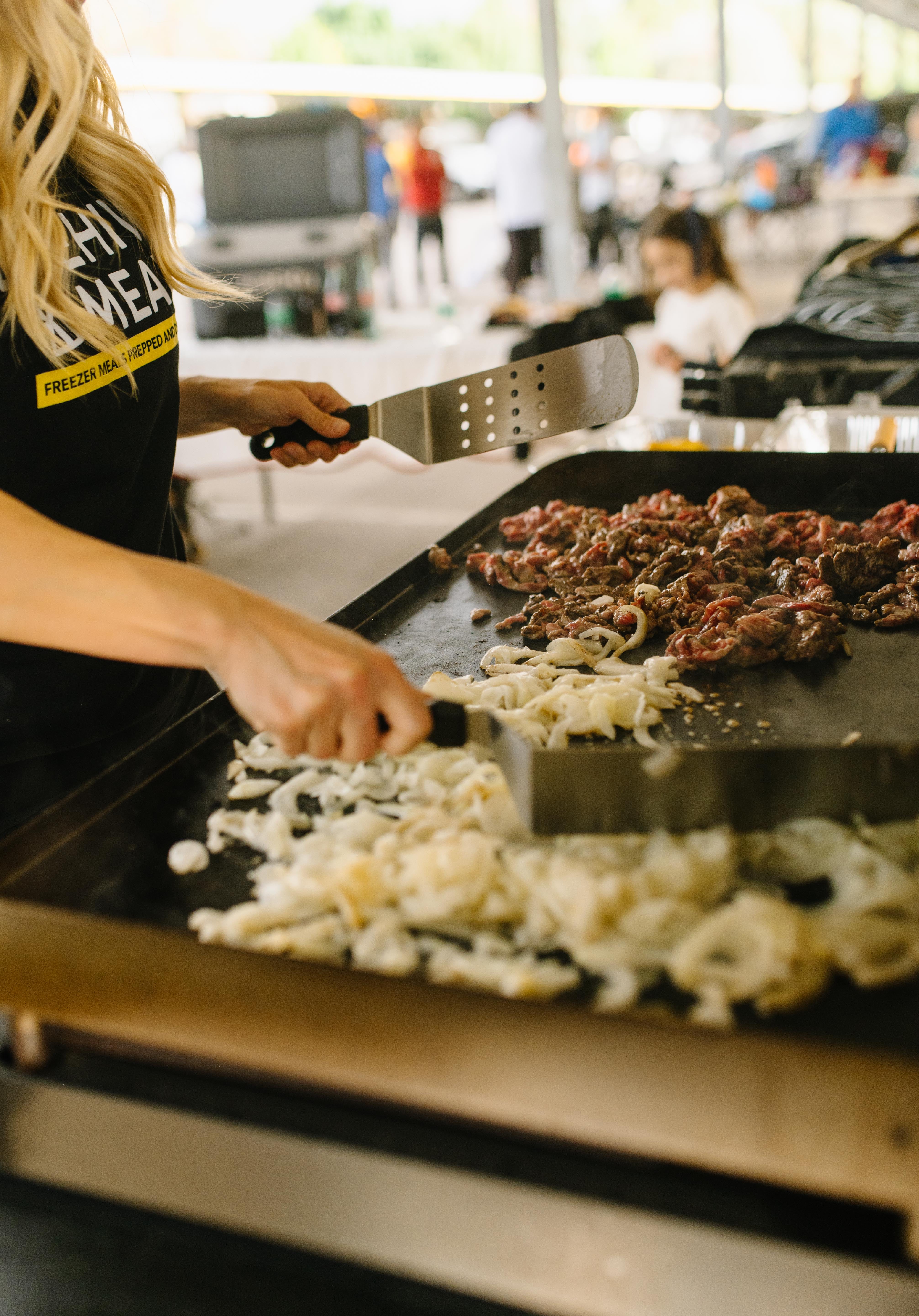 Women cooking on a flat top grill with food and utensils in an outdoor setting.