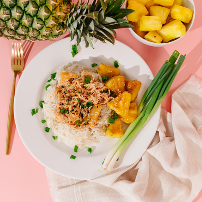 A styled overhead food shot of a white plate with white rice topped with shredded chicken in a glossy orange sauce, garnished with chopped chives, served with pineapple chunks and green onions on a pink background. A whole pineapple, a bowl of pineapple pieces, a gold fork, and a beige cloth are also visible.