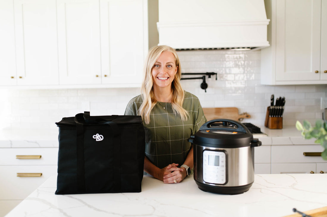 Allyse, founder of Beehive Meals, stands in a bright white kitchen behind a marble counter, presenting a large black insulated tote bag next to a slow cooker. 