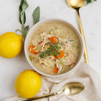 Bowl of Beehive Meals Lemon Chicken Orzo Soup with lemons and herbs on a marble surface with gold spoons.