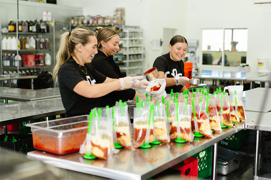 Three women working in the Beehive Melas prep kitchen preparing food packages.