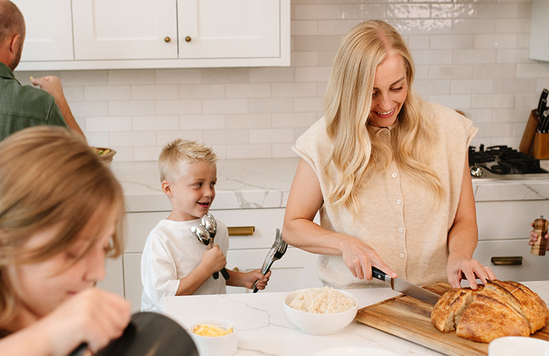 A woman in a beige sleeveless top slices a loaf of bread on a wooden cutting board in a bright, modern kitchen, with a young boy holding metal utensils nearby and another person partially visible in the foreground.