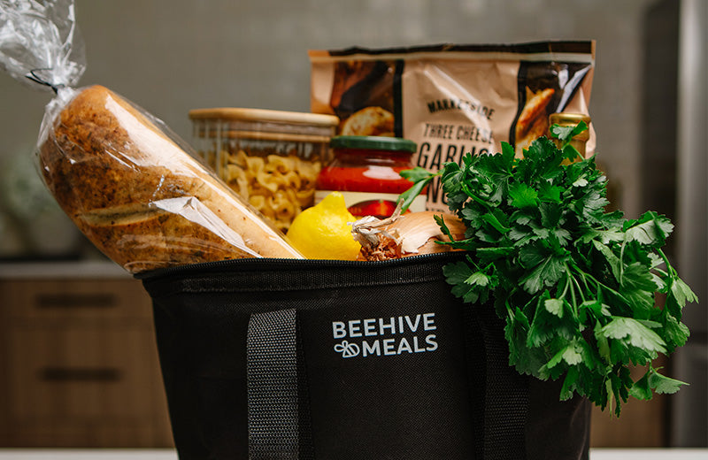 A black Beehive Meals insulated food bag filled with groceries, including a loaf of bread, pasta, a bag of three cheese garlic noodles, a lemon, and a bunch of fresh parsley, set against a blurred kitchen background.