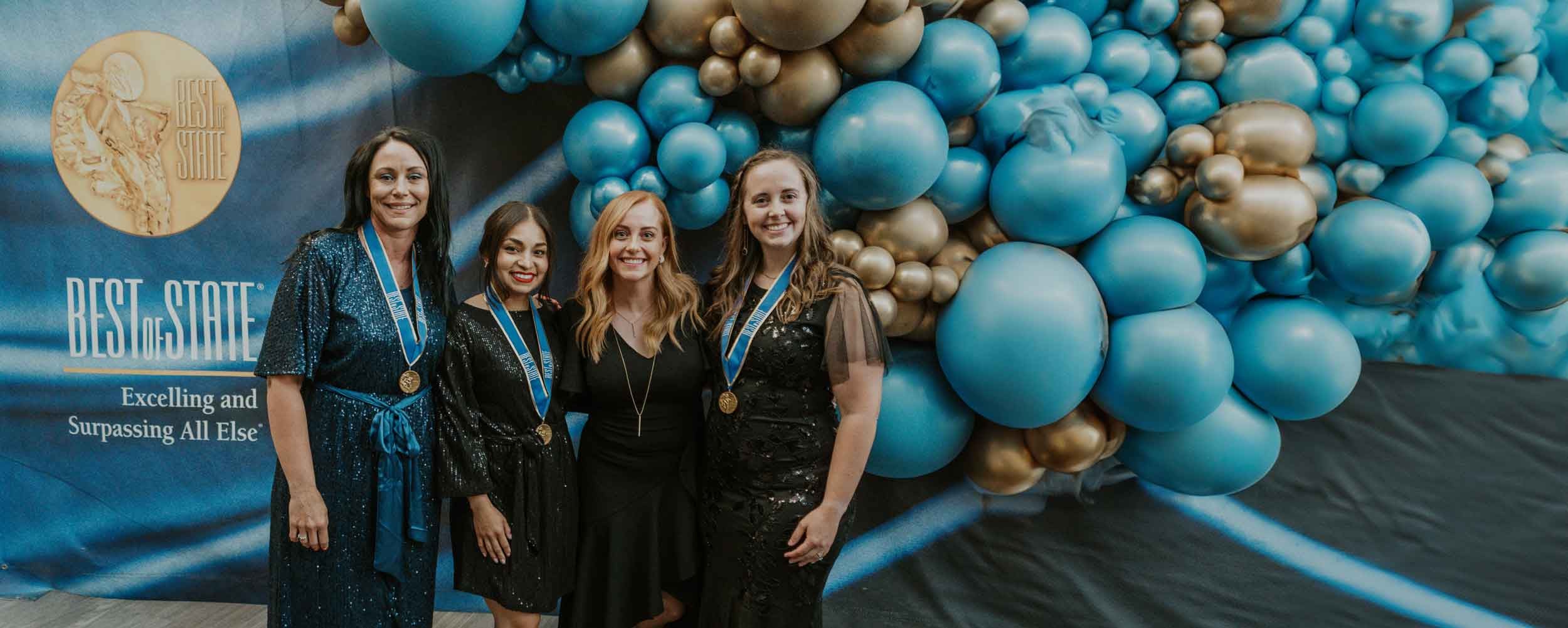 Four women posing with three Best of State awards in front of a balloon arch.