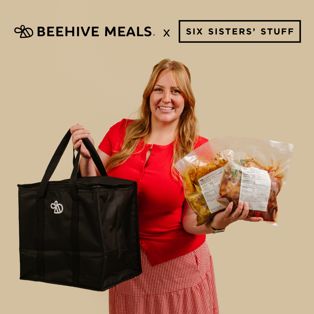 A woman in a red top and pink gingham skirt holds a large black tote bag branded Beehive Meals in one hand and a clear bag of food in the other, against a solid beige background with Beehive Meals and Six Sisters' Stuff branding at the top.